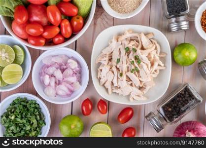 Boiled chicken cut into pieces in a white dish, along with lemons, chilies, lemongrass, red onions, chopped onions, tomatoes, sesame, roasted rice, and pepper on a wooden table.