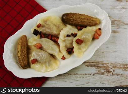Boiled dumplings with meat topped with fried bacon with pickled cucumbers on white platter on an old wooden table. Traditional dish of Eastern Europe. Flat, horizontal view from the top.