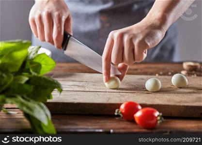Boiled quail eggs cut female hands on a wooden board on a table with tomatoes and spinach. Step by step preparation. On a wooden table, the hands of a woman cut quail eggs on an old wooden board. Step by step salad preparation