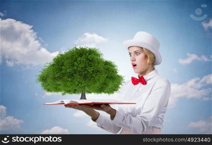 Book that blow up your imagination. Young woman in white hat with opened book in hands and green tree on pages