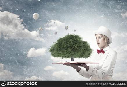 Book that blow up your imagination. Young woman in white hat with opened book in hands and green tree on pages