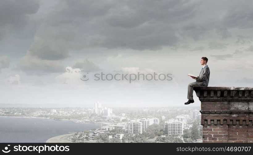 Books broaden your mind. Young businessman sitting on building top and reading book