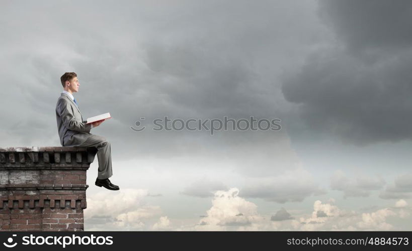 Books broaden your mind. Young businessman sitting on building top and reading book