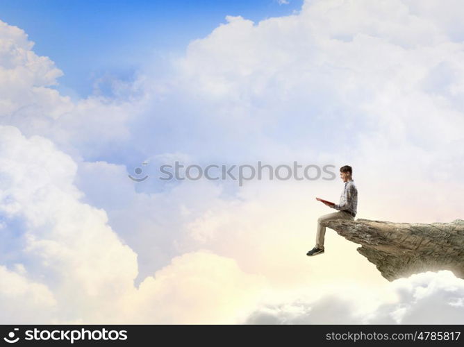 Books let you rise above the rest. Young man in casual sitting on rock top with book in hands