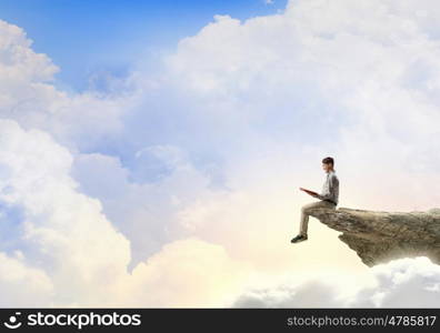 Books let you rise above the rest. Young man in casual sitting on rock top with book in hands