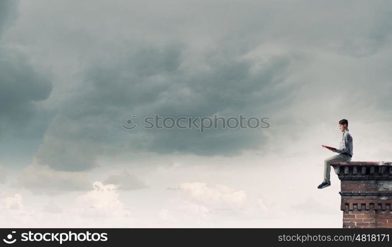 Books let you rise above the rest. Young man in casual sitting on building top with book in hands