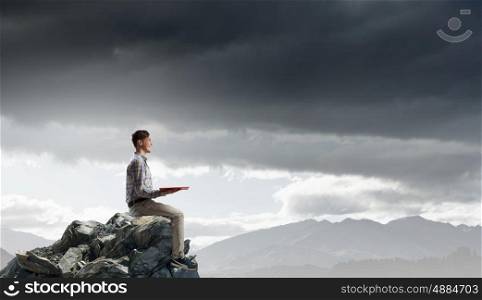 Books let you rise above the rest. Young man in casual sitting on rock top with book in hands