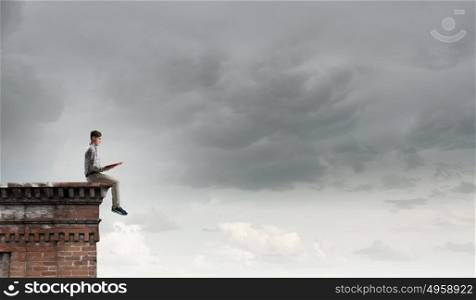 Books let you rise above the rest. Young man in casual sitting on building top with book in hands