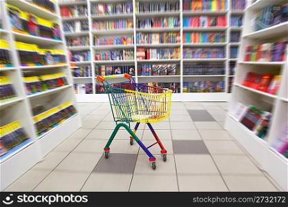 Bookshop interior. Before racks with books there is bright cart for purchases. Blurred.