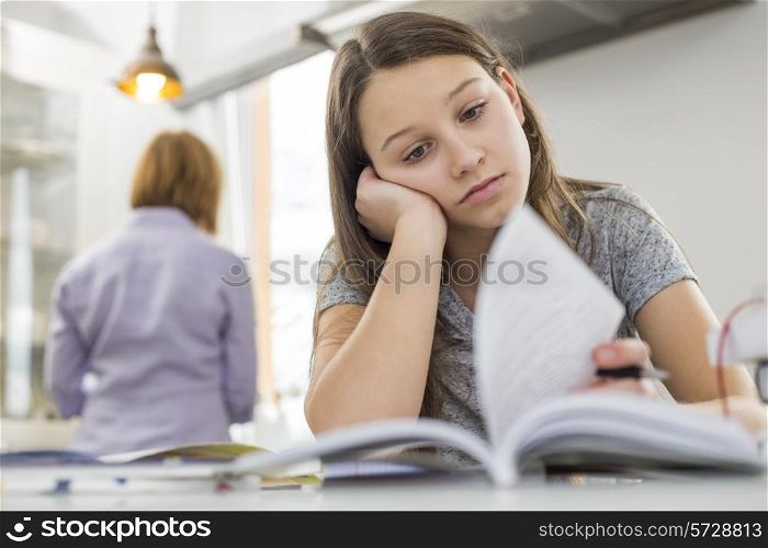 Bored girl studying at table with mother standing in background