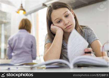 Bored girl studying at table with mother standing in background
