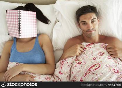 Bored Looking Man Lying In Bed Next To Woman Reading Book