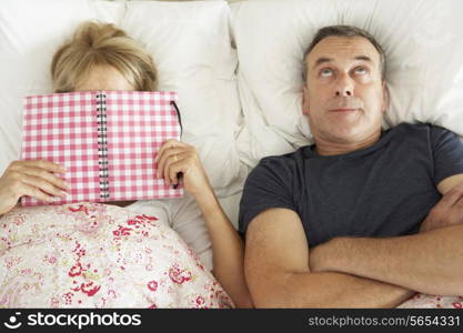 Bored Looking Senior Man Lying In Bed Next To Senior Woman Reading Book