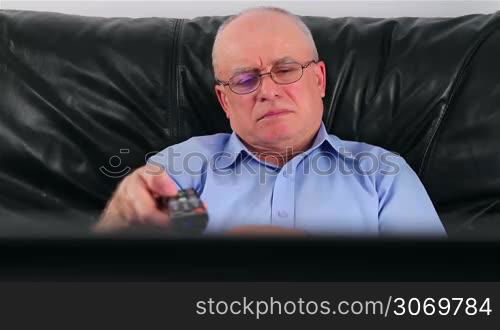 Bored senior man with remote control watching TV on sofa at home
