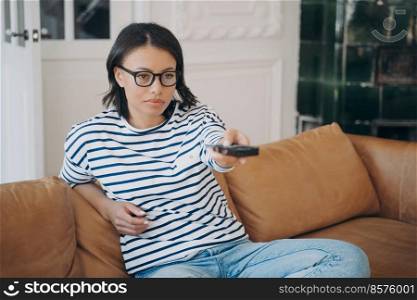 Bored young caucasian woman sitting on leather couch and watching tv. Lady in glasses is holding remote controller and shifts channels. Boring movie or show. Leisure and relaxation at home.. Bored young caucasian woman holding remote controller and watching boring movie or tv show.