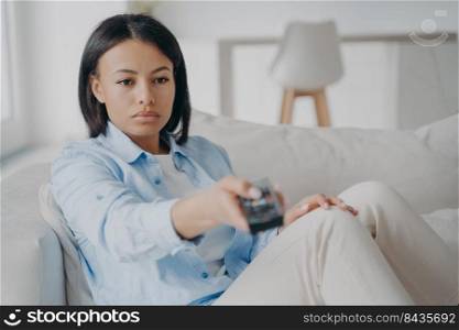 Bored young female holding tv remote control change channels lost in thoughts sitting alone on couch at home. Tired woman spend free time watching television set after hard work day.. Bored female holding tv remote control change channels, watching television sitting on couch at home