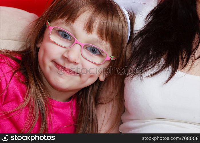 Boredom, vision, eye problems concept. Young toddler girl wearing glasses sitting on sofa next to her mother indoor. Young toddler girl in glasses sitting on sofa