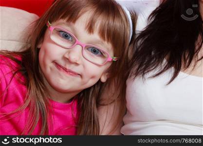 Boredom, vision, eye problems concept. Young toddler girl wearing glasses sitting on sofa next to her mother indoor. Young toddler girl in glasses sitting on sofa