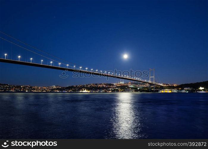 Bosphorus Bridge illuminated by lights and moon at night. Bridge over the Bosphorus to the coast with lighted houses under a bright moon. Istanbul, Turkey.. Bosphorus Bridge illuminated by lights and moon at night