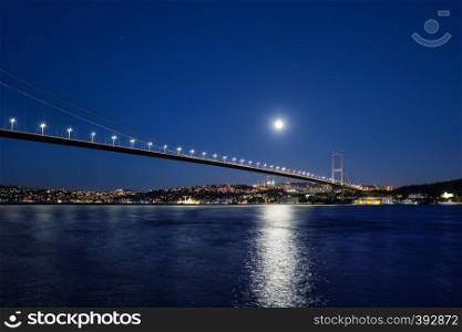 Bosphorus Bridge illuminated by lights and moon at night. Bridge over the Bosphorus to the coast with lighted houses under a bright moon. Istanbul, Turkey.. Bosphorus Bridge illuminated by lights and moon at night