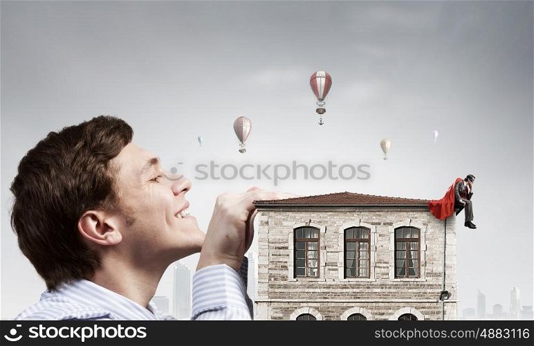 Boss peeping from under table. Bored businessman with red cape sitting on the roof of building