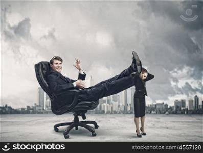 Bossy businessman. Young businessman sitting gin chair with legs on colleagues shoulders