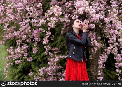 botanical garden, young girl, sweet woman, leather jacket, flowering trees, flowers on a tree, sakura, pink flowers. A young girl in a black jacket and a red dress stands next to sakura and sniffs her