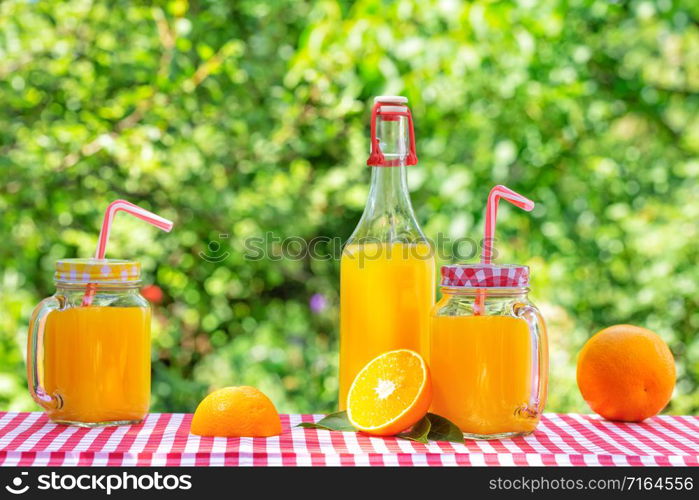 Bottle and cans of orange juice with oranges on a checkered tablecloth. Natural green background. Eco-friendly Glassware. Bottle and cans of orange juice with oranges on checkered tablecloth