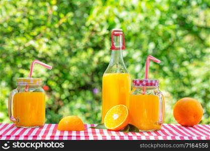 Bottle and cans of orange juice with oranges on a checkered tablecloth. Natural green background. Eco-friendly Glassware. Bottle and cans of orange juice with oranges on checkered tablecloth