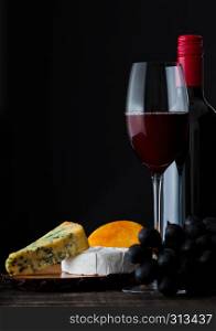 Bottle and glass of red wine with cheese selection with grapes on black background