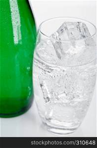 Bottle and glass with healthy sparkling water with ice cubes on white