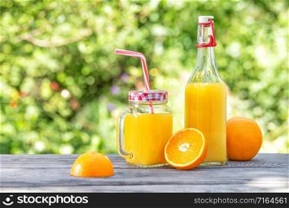 Bottle and jar with orange juice and oranges on a wooden table. Green natural background. Summer still life. Bottle and jar with orange juice and oranges on wooden table