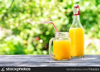 Bottle and jar with orange juice on a wooden table. Green natural background. Summer still life. Bottle and jar with orange juice on a wooden table