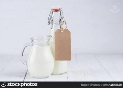 Bottle of milk and jar of milk with blank cardboard label on white wooden table background