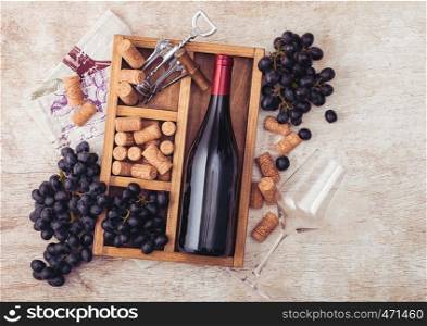 Bottle of red wine and empty glass with dark grapes with corks and opener inside vintage wooden box on light wooden background with linen towel
