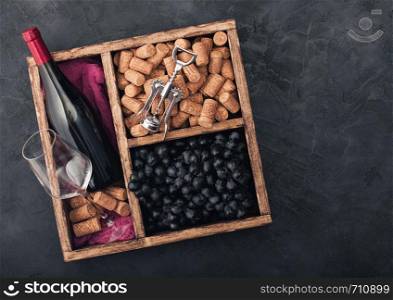 Bottle of red wine and empty glass with dark grapes with corks and corkscrew inside vintage wooden box on black stone background with red cloth. Space for text