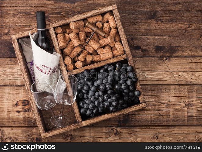 Bottle of red wine and empty glass with dark grapes with corks and opener inside vintage wooden box on grunge wooden background with linen towel.