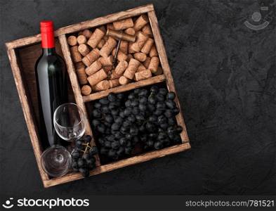Bottle of red wine and empty glasses with dark grapes with corks and opener inside vintage wooden box on black stone background.