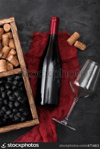 Bottle of red wine on red cloth with empty glass and dark grapes with corks and corkscrew inside vintage wooden box on dark wooden background.
