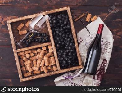 Bottle of red wine on wood with empty glass with dark grapes with corks and corkscrew inside vintage wooden box on dark wooden background with linen towel.