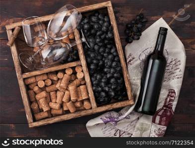 Bottle of red wine on wood with empty glasses with dark grapes with corks and corkscrew inside vintage wooden box on dark wooden background with linen towel.
