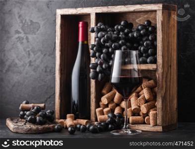 Bottle of red wine with dark grapes and corks inside vintage wooden box on black stone background. Elegant wine glass with corkscrew on black board.