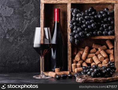 Bottle of red wine with dark grapes and corks inside vintage wooden box on black stone background. Elegant wine glass with corkscrew on black board. Space for text
