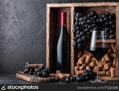 Bottle of red wine with dark grapes and corks inside vintage wooden box on black stone background. Elegant wine glass with corkscrew on black board.