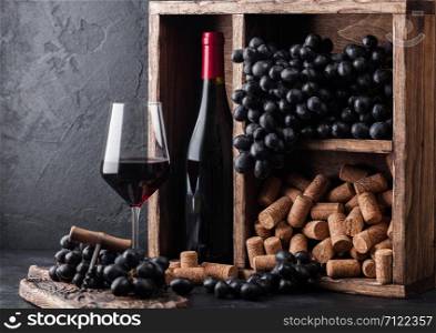 Bottle of red wine with dark grapes and corks inside vintage wooden box on black stone background. Elegant wine glass with corkscrew on black board. Wine still life concept