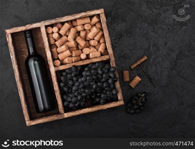 Bottle of red wine with dark grapes with corks and opener inside vintage wooden box on black background.