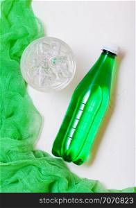 Bottle of sparkling mineral water with glass of ice and green cloth on white background