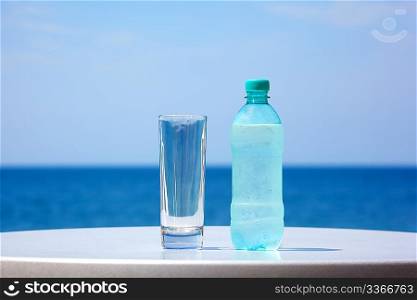Bottle of water and glass on table under open sky on background of sea.