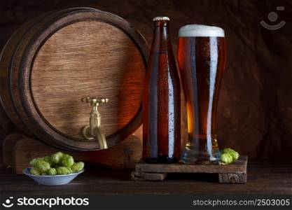Bottled and unbottled beer glass with barrel and fresh hops for brewing on wooden table still-life