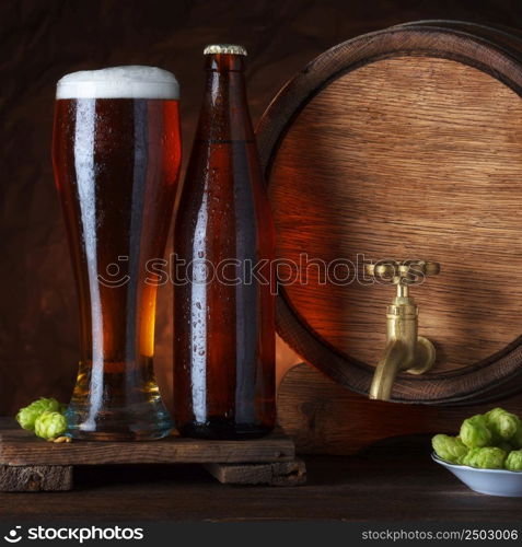 Bottled and unbottled beer glass with barrel and fresh hops for brewing on wooden table still-life